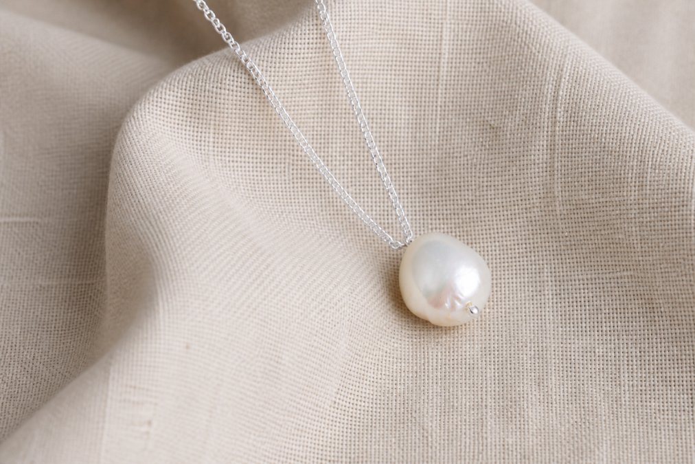 Close-up of a delicate baroque pearl suspended on a fine sterling silver chain against natural linen fabric, showing minimalist elegance and timeless design in soft daylight
