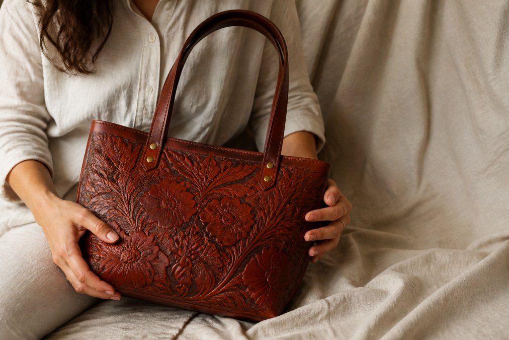 Close-up of a woman's hands holding a hand-tooled floral vegan leather tote bag, detailed embossing visible against a neutral linen background, soft natural window light