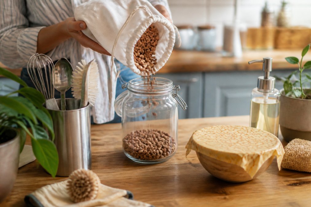 Hand pouring dried beans from a cloth bag into a glass jar on a wooden kitchen counter with eco-friendly supplies.