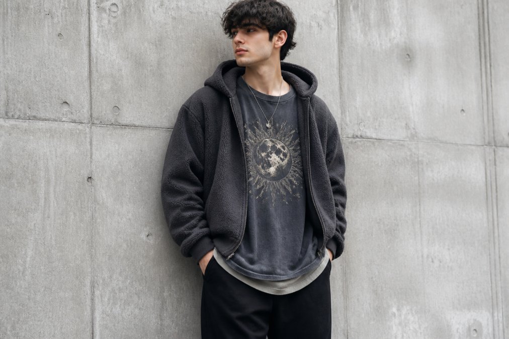 A young adult wearing an oversized vintage-washed graphic tee layered under a boxy fleece hoodie, standing against a plain concrete wall in soft daylight, relaxed pose, neutral color palette