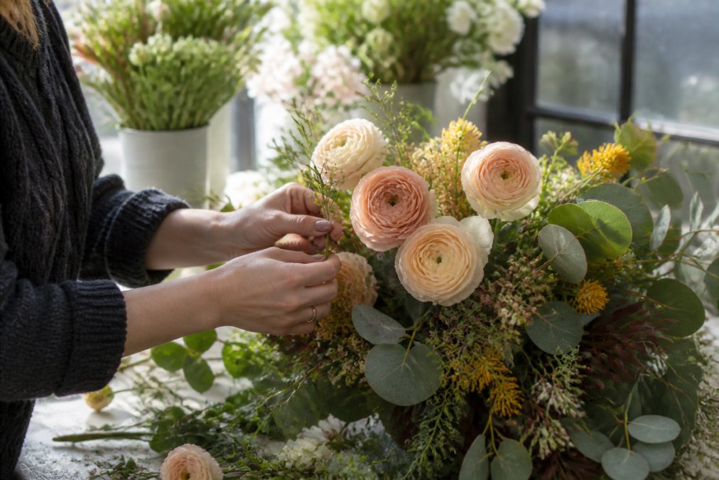 Florist's hands arranging fresh seasonal blooms including ranunculus, eucalyptus greenery, and native Australian flowers with morning dew glistening on petals in natural window light
