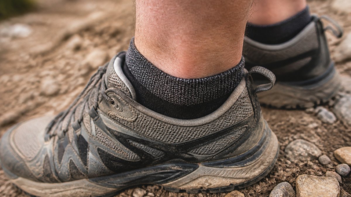Close-up of a trail runner's feet on a dusty rocky path, showing a low-cut sock with a visible debris-blocking cuff sitting above a trail running shoe collar, with small rocks and grit scattered nearby