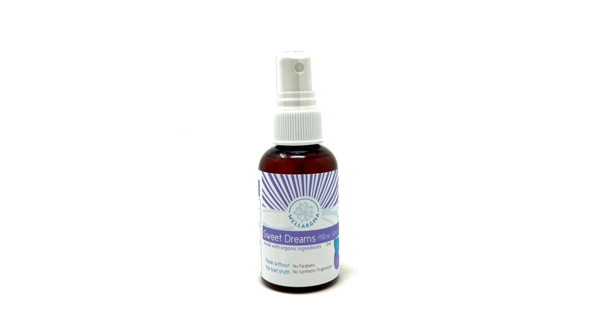 Close-up of Wellaroma's Sweet Dreams Pillow Spray bottle and Magnesium Bedtime Spray next to dried lavender flowers and a white pillow on a dark wooden nightstand, with soft warm lighting from a bedside lamp