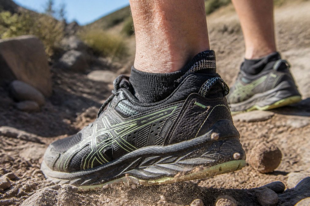 Close-up of a trail runner's foot stepping onto rocky mountain terrain, showing a technical sock's reinforced cuff and debris-resistant material deflecting small stones and dirt particles
