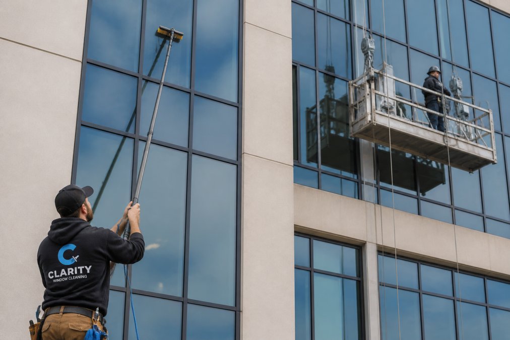 A commercial window cleaner using a long carbon fiber water fed pole from the ground to clean glass on a multi-story office building, with a swing stage suspended platform visible on the adjacent facade