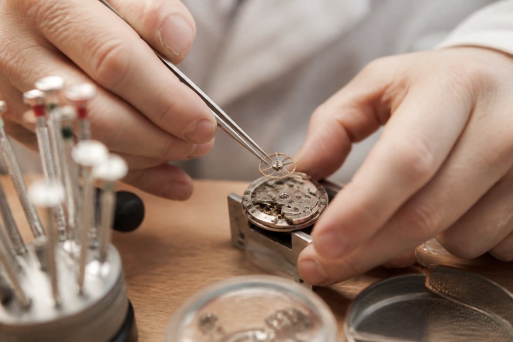 Close-up of a watchmaker's hands in a well-lit workshop, carefully placing a tiny balance wheel into a mechanical movement using fine steel tweezers