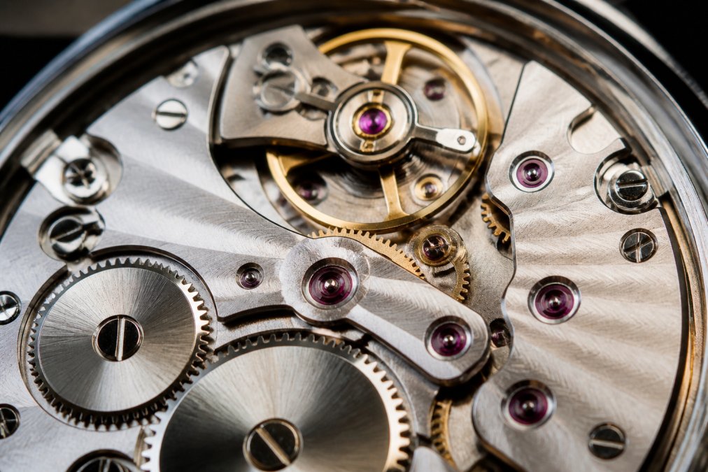 Close-up of a watch movement showing jeweled bearings, escapement, and hand-finished components with warm lighting highlighting intricate mechanical details and polished surfaces