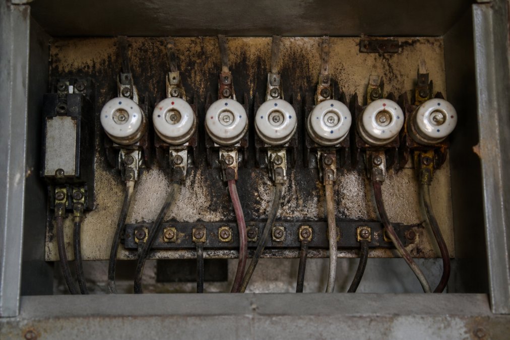 Close-up of an older switchboard with ceramic fuses, worn metal components, and discoloured backing showing age and corrosion in a dimly lit residential setting