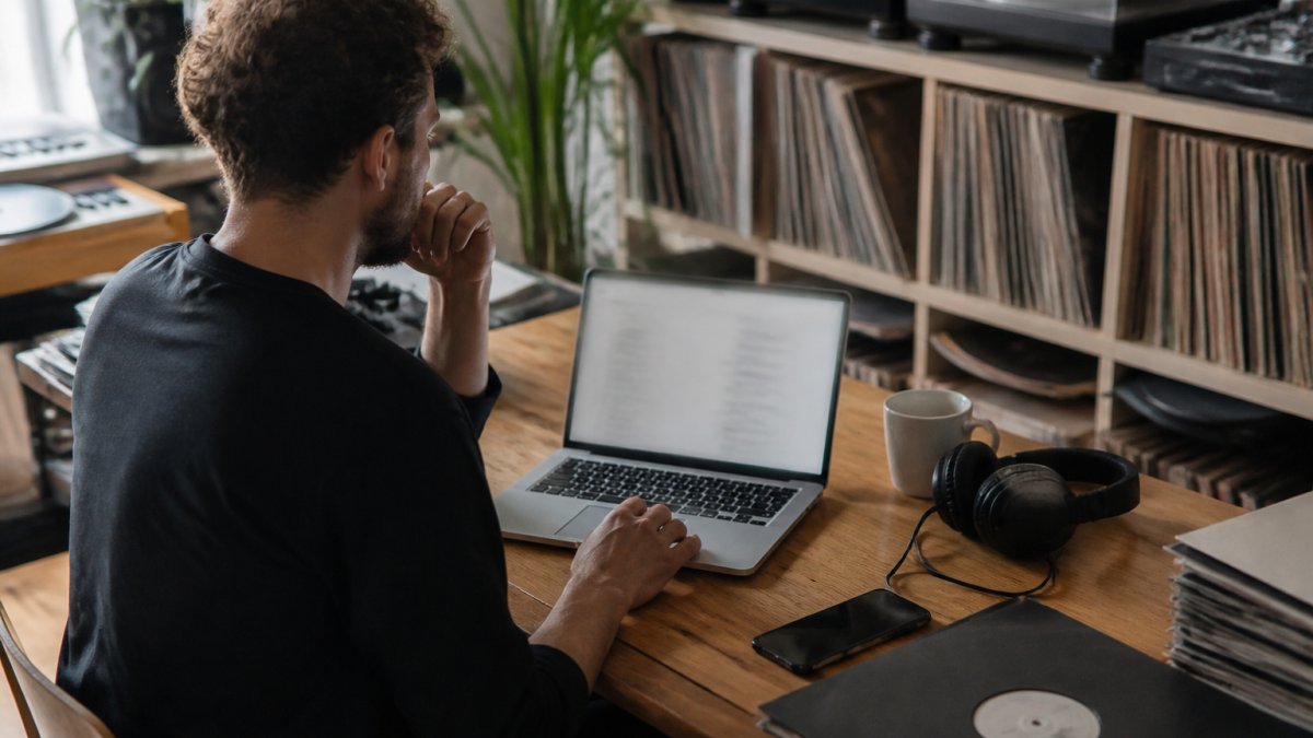 Music producer at home studio reviewing sync pitch emails on laptop screen with headphones on desk and vinyl records on shelves