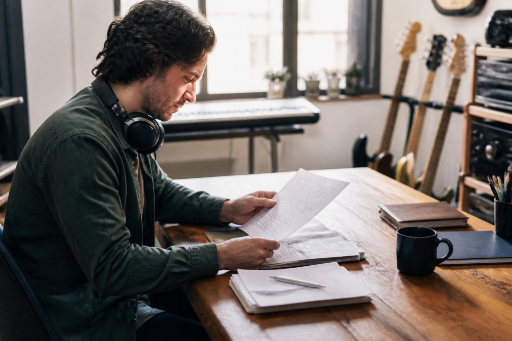 Music producer reviewing licensing documents and notes on a laptop displaying music production software, with headphones around neck on a wooden desk in natural daylight