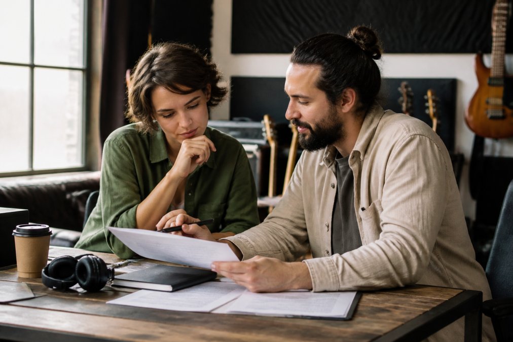 Two creative professionals collaborating over a laptop in a studio setting, reviewing licensing agreements and music project details with natural lighting
