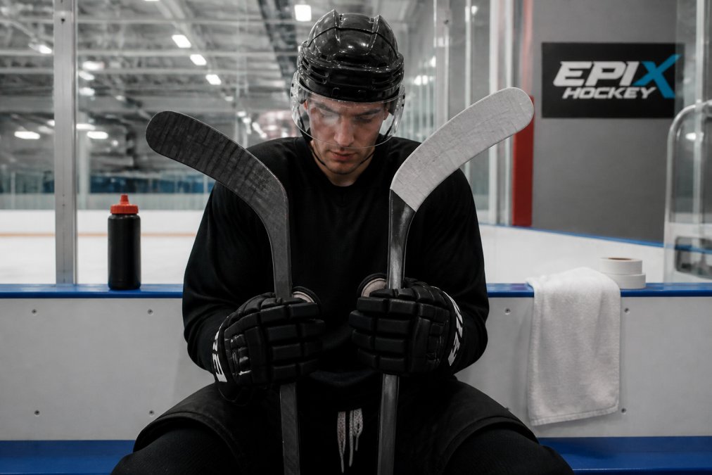 A hockey player in full gear holding two sticks side by side on the bench, comparing blade shapes and tape jobs under rink lighting