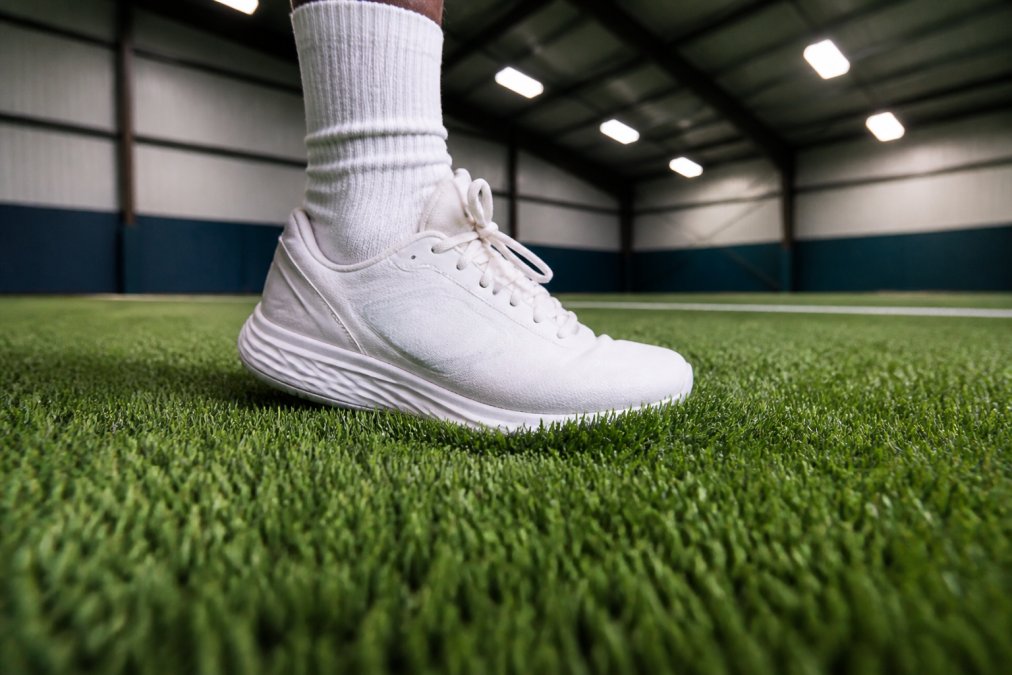 Close-up of athlete's foot striking padded synthetic turf surface in indoor gym, showing cushioning compression and fiber contact detail with focused lighting