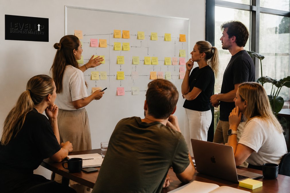 Service business team gathered around whiteboard in modern office, reviewing workflow diagrams with sticky notes marking process steps, handoffs, and delays in natural daylight