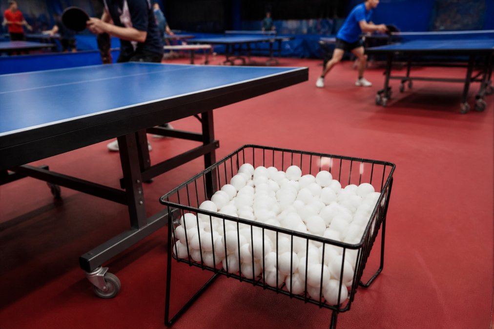 A wire basket full of white ping pong balls sitting on the floor beside a table tennis table in a club hall