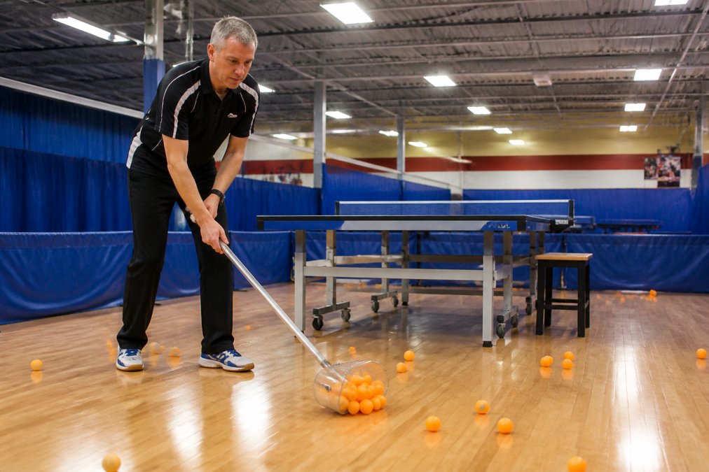 A coach using a handheld ball collector tool to scoop scattered orange table tennis balls off a hardwood floor, with a full-size table tennis table and club banners visible in the background under bright gymnasium lighting