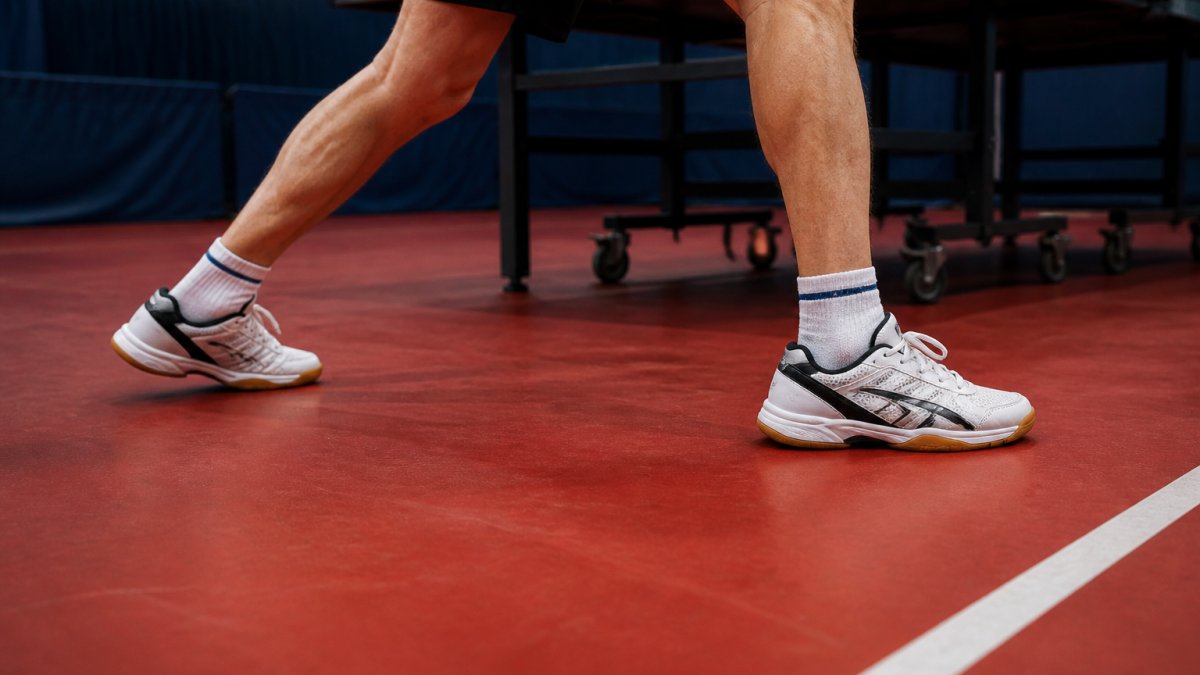 Senior player's feet in table tennis shoes positioned on indoor court during lateral movement, demonstrating proper stance and foot positioning with visible shoe support