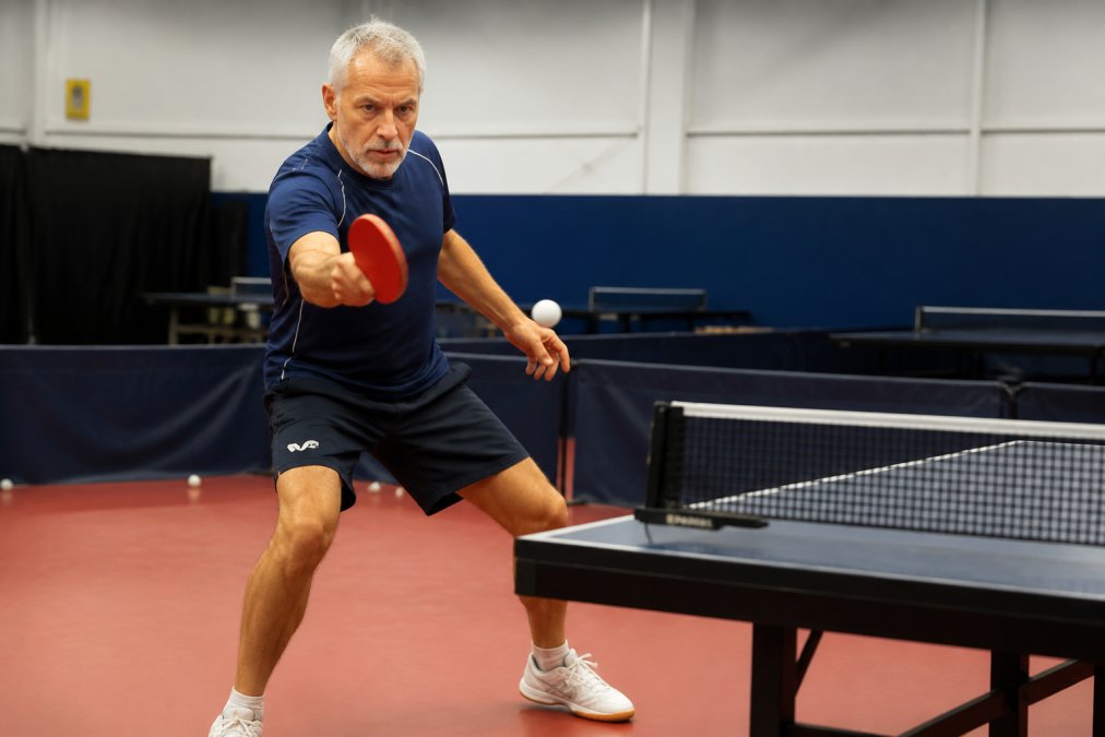 Active senior player in athletic wear executing a forehand stroke at a table tennis table, demonstrating proper form and stance with engaged core and bent knees