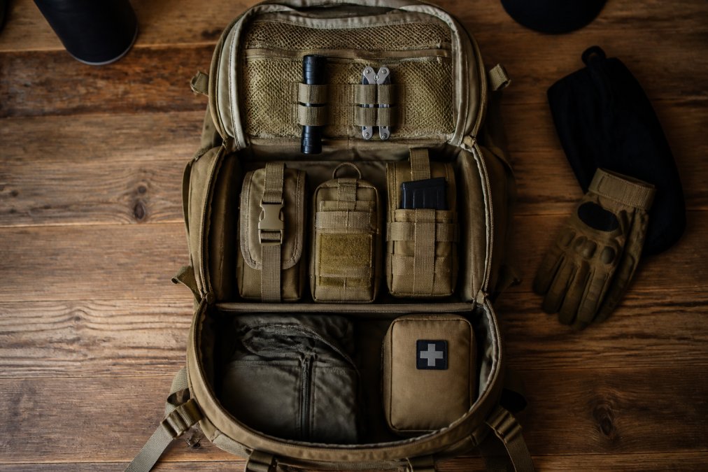 Overhead shot of an open tactical backpack on a wooden table, gear sorted into three distinct zones: top layer flashlight and multi-tool, middle layer with modular pouches, bottom layer with folded jacket and first-aid kit, warm natural lighting