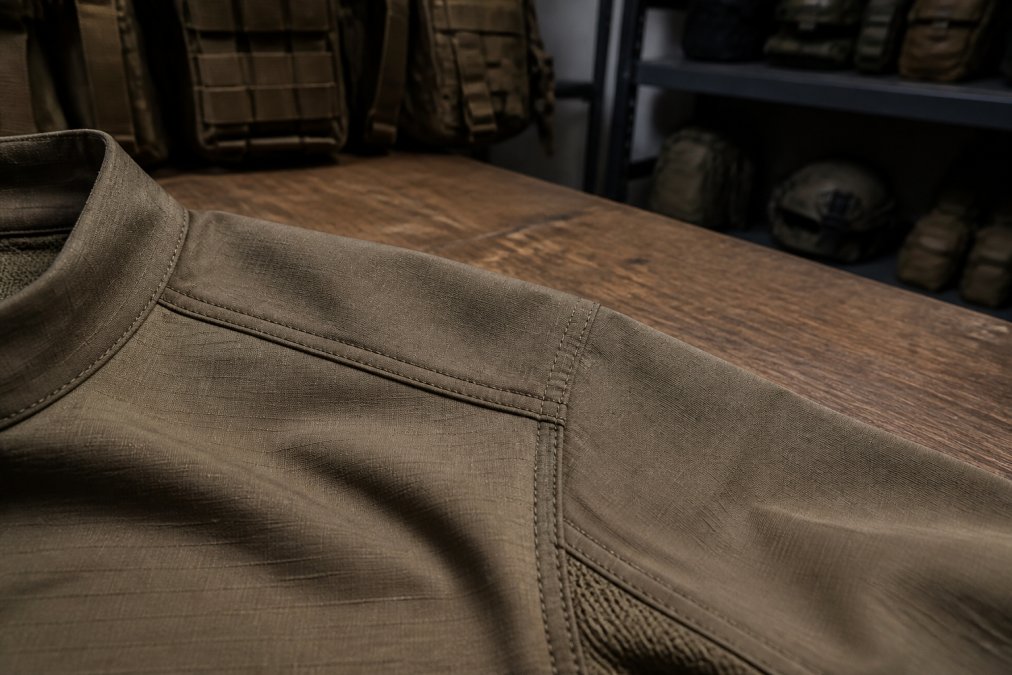 Close-up of a tactical shirt shoulder seam showing double-stitched reinforced thread, resting on a wooden workbench in a gear room with soft overhead lighting