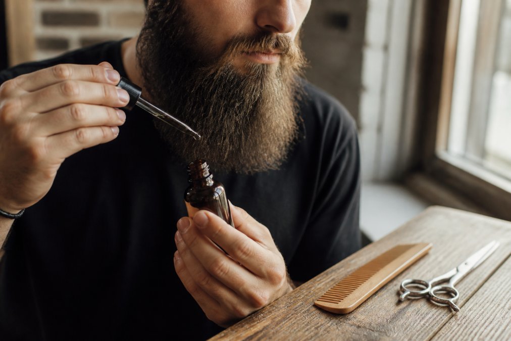 Gros plan sur les mains d'un homme appliquant de l'huile à barbe sur une barbe longue et soignée, un peigne et des ciseaux posés sur une surface en bois à côté, lumière naturelle venant d'une fenêtre