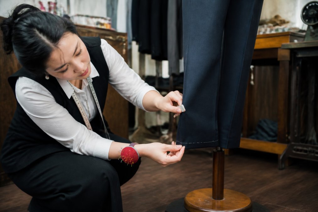 A skilled tailor measuring the hem of dress trousers on a fitting dummy in a professional alteration studio, showing precision work with fabric and chalk marking