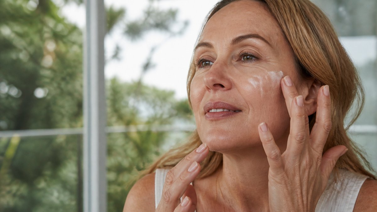 Close-up of mature woman's face with glowing, hydrated skin as she gently applies moisturiser to her cheek and jawline with fingertips