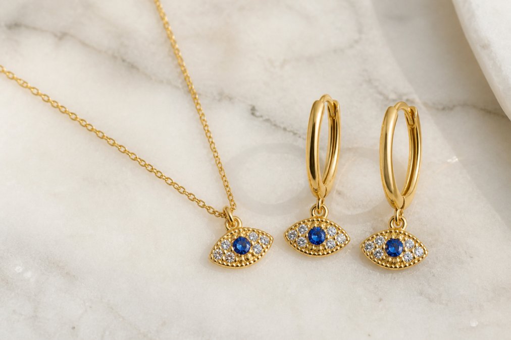 Close-up of a gold-plated evil eye necklace and huggie hoop earrings laid flat on a light marble surface, showing vibrant blue enamel evil eye detail and shimmering zirconia stones in warm natural light