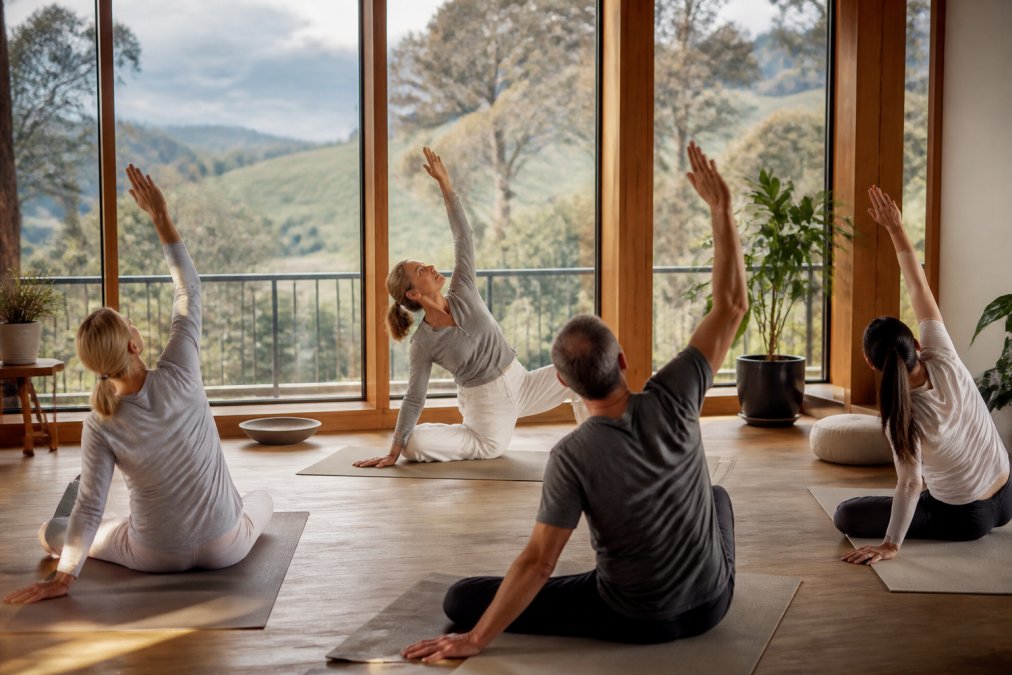 Small group of guests in gentle yoga pose in light-filled room overlooking natural landscape during morning session