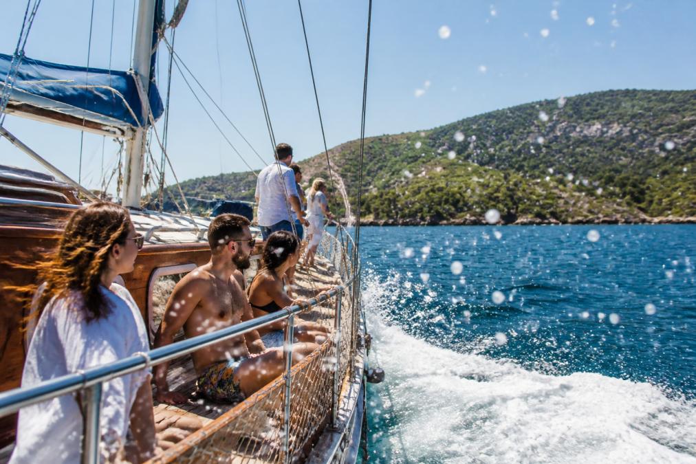Turkuaz suların üzerinde yelkenli teknede turist grubu, güneşli bir günde kıyı manzarasıyla birlikte, dalgaların teknede sıçrayan su damlaları görülüyor