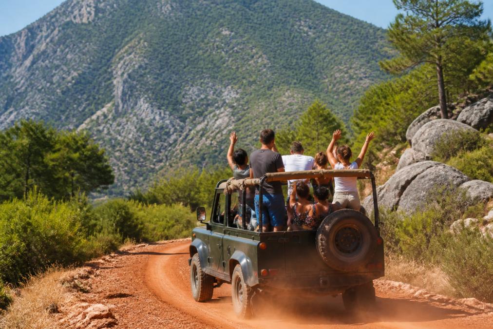 Dağlık arazi üzerinde jeep safari aracında heyecanlı gezginler, yeşil orman ve doğal manzara arka planında, araç kırmızı toprak yolunda hareket ederken