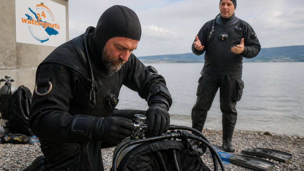 Erfahrener Taucher in Trockentauchanzug prüft Ausrüstung am Ufer des Bodensees, Tauchlehrer steht im Hintergrund und erklärt die Sicherheitsschritte