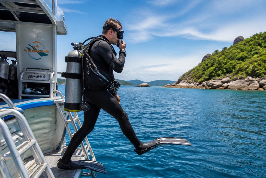Ein Taucher in voller Ausrüstung am Rand eines Bootes, der vor einer tropischen Küste ins Wasser einsteigt, mit Tauchcomputer und Atemregler sichtbar