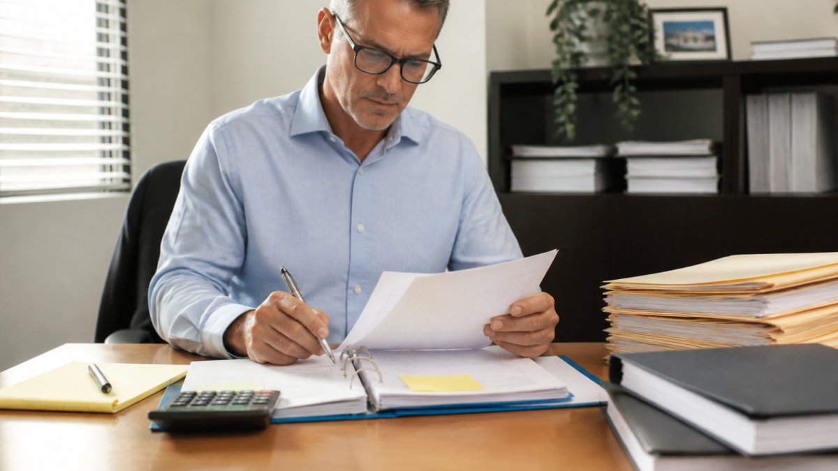 Business professional sitting at organized desk reviewing financial documents and organized files with calculator and notepad, natural office lighting