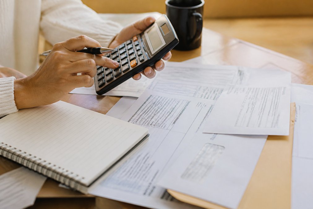 Close-up of hands holding calculator and reviewing financial statements with IRS correspondence on desk, notepad visible, warm office lighting