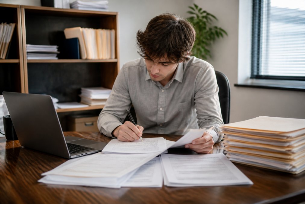 Young accounting student studying tax documents and IRS forms at a desk with organized file folders and laptop displaying tax software interface, natural office lighting