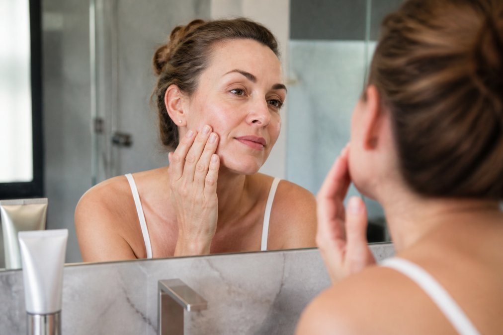 A woman in her 40s examining her skin in a bathroom mirror, with a tube of post-peel moisturizer and sunscreen on the counter, soft natural lighting