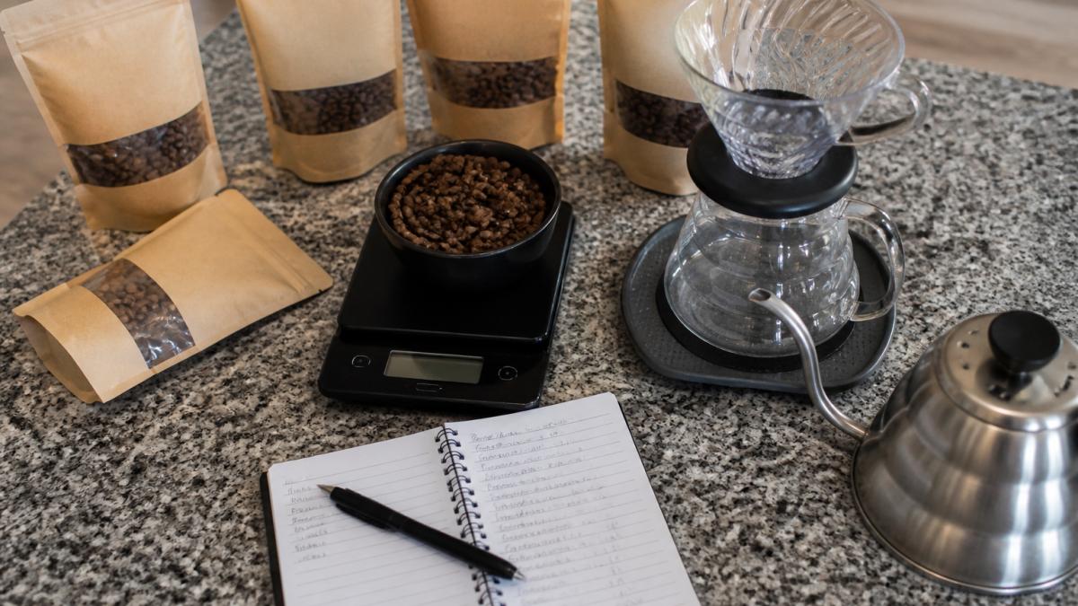 Overhead shot of a kitchen counter with a digital scale, several 2oz coffee sample bags, a pour-over dripper, and a small notebook with tasting notes written in pen