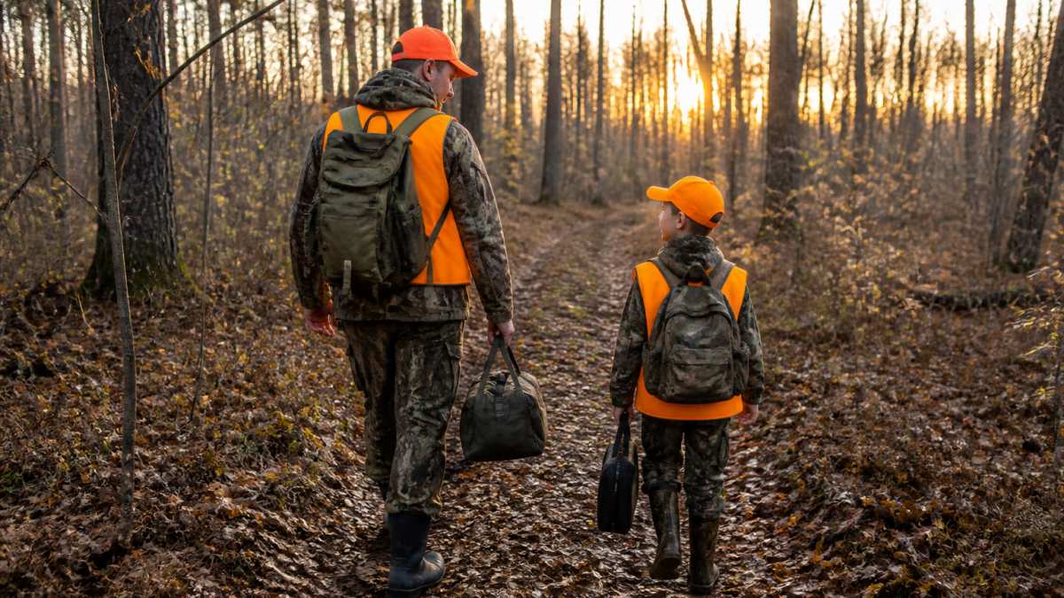 A parent and young child in blaze orange gear walking together through a wooded trail at dawn, both carrying gear and looking engaged