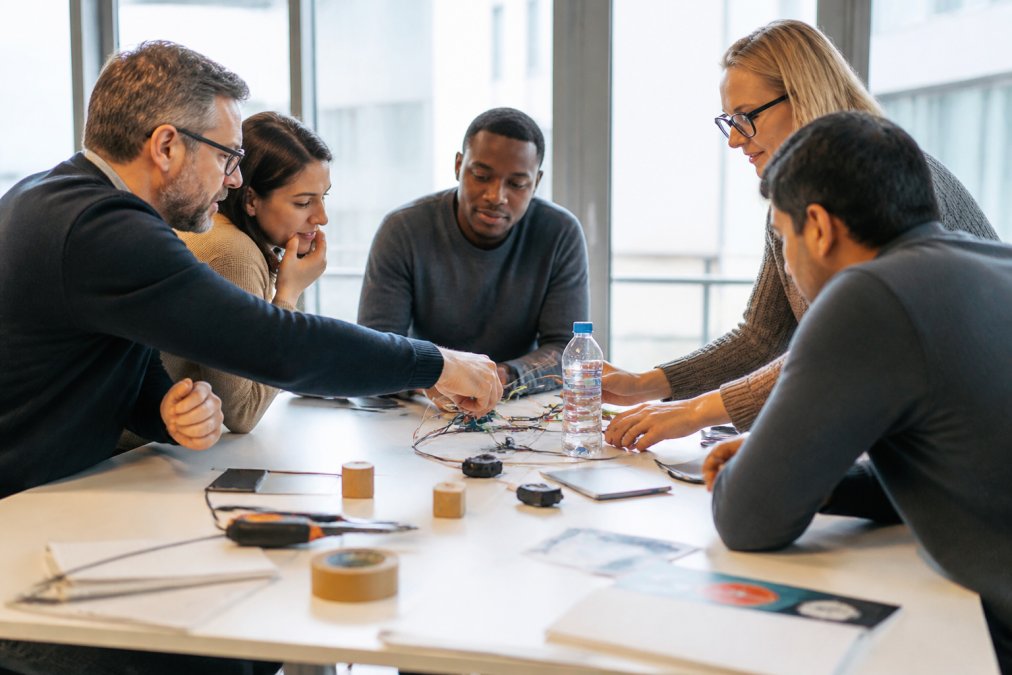 A mixed group of employees from different departments collaborating around a large table, working on a hands-on problem-solving activity with materials and notes visible, in a bright modern workspace