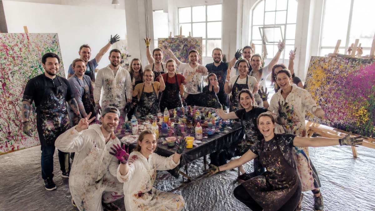 Eine Gruppe von etwa 25 Personen beim gemeinsamen Action Painting Workshop, mit bunten Farben auf großen Leinwänden, lachende Gesichter und kreatives Durcheinander in einer inspirierenden Studio-Atmosphäre mit natürlichem Licht