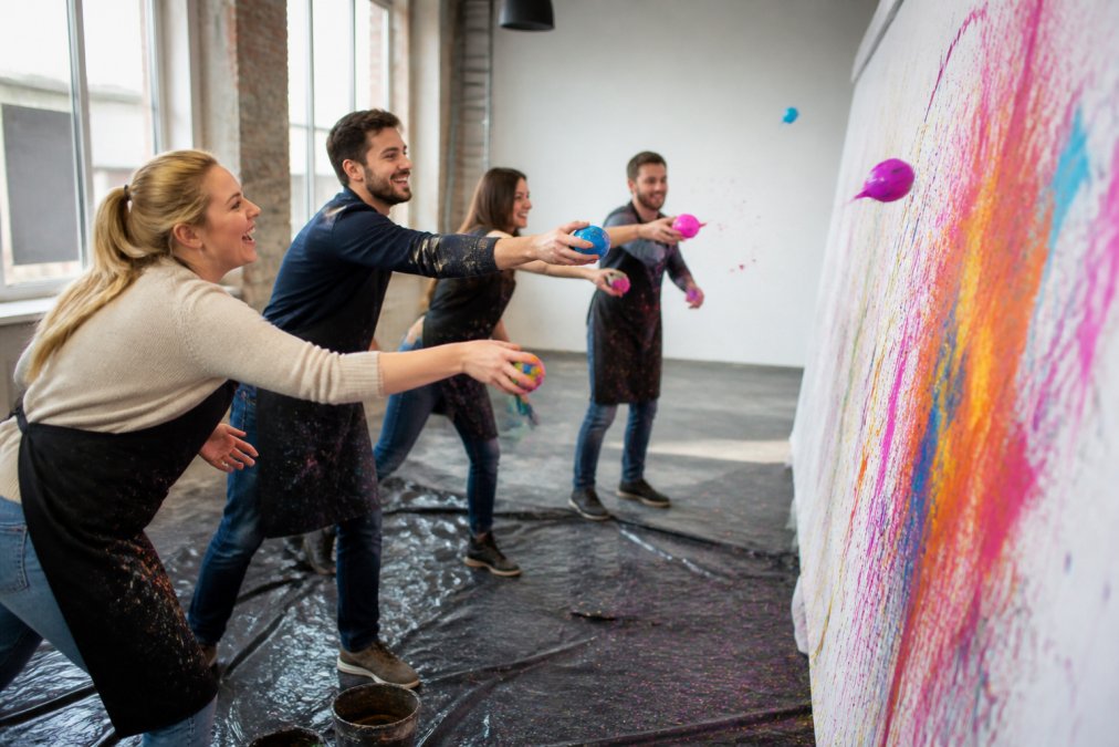 A group of colleagues in casual clothing throwing paint-filled balloons at a large canvas in a bright, spacious studio, laughing and working together, colorful splashes on the canvas and floor coverings