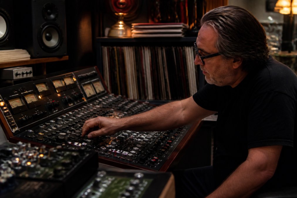 Professional mastering engineer adjusting analog outboard gear on a mixing console in an acoustically treated studio, with monitor speakers and vinyl records visible in the background under warm studio lighting
