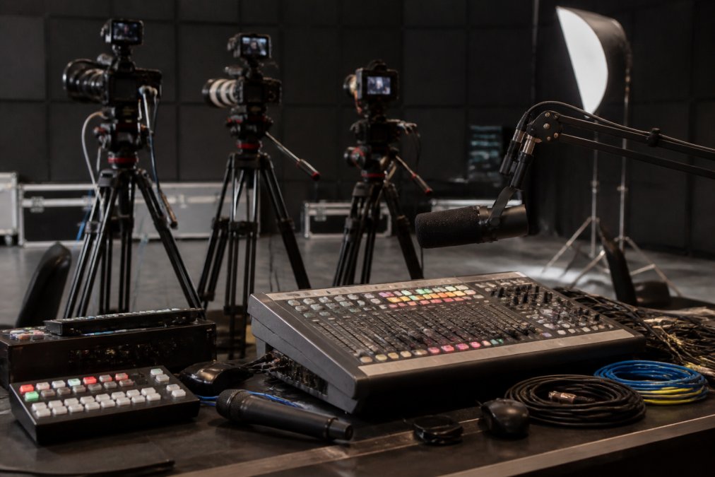 Professional streaming setup with multi-camera rig mounted on tripods, mixing console with faders and buttons, professional microphone on boom arm, LED lighting panels, and network cables organized on a production desk in a broadcast studio with dark walls