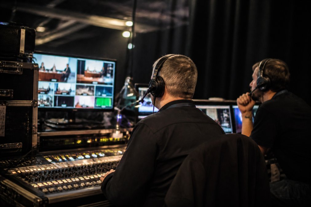 Technical director and on-site engineer wearing headsets, monitoring live stream on three large screens displaying video feeds, waveforms, and performance metrics, with control panels and mixing console in foreground, in professional broadcast control room with dim lighting