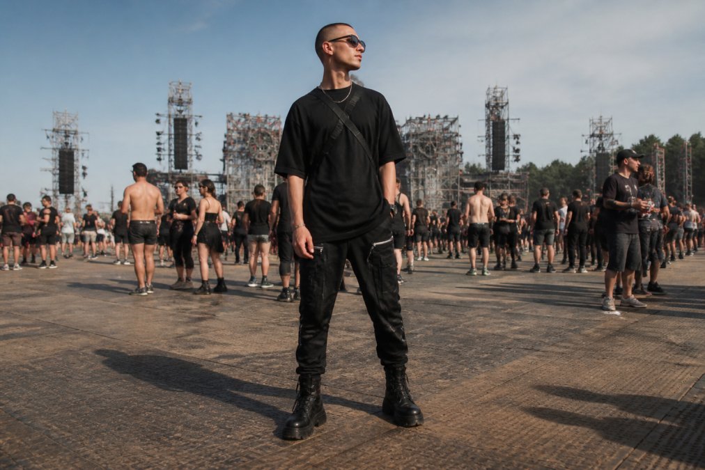 Person wearing a complete techno outfit on a festival ground: black oversized shirt, utility pants with reflective details, platform boots, and minimal accessories, posed confidently during daytime at an outdoor venue
