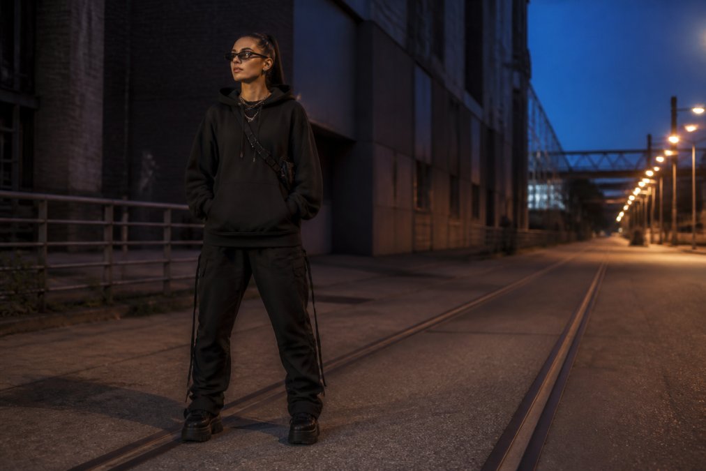 Person iført komplet techno-streetwear-outfit med oversized hættetrøje i sort, mørke bukser, platformssneakers og minimalistisk rave-tilbehør på en urban gade med industriel baggrund og aftenlys