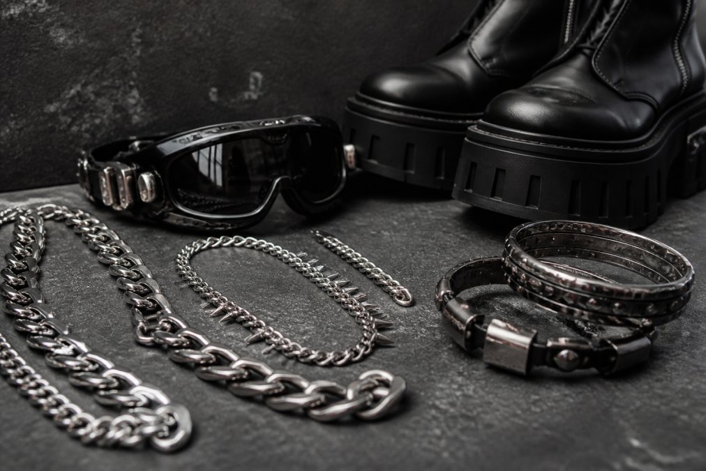 Nærbillede af forskelligt techno-tilbehør, herunder sølvkæder, futuristiske goggles med mørke glas, detaljer af plateausko og metalarmbånd arrangeret på en mørk tekstur med industrielt lys