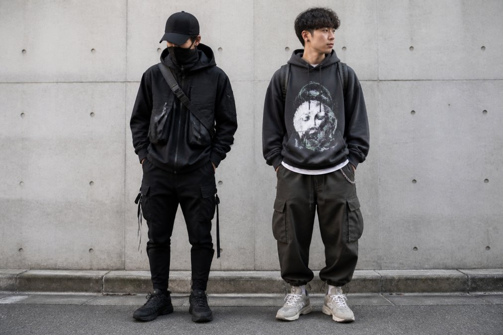 Two men standing side by side against an urban concrete wall: one wearing a sleek black technical jacket with utility straps and tapered trousers, the other in a baggy graphic hoodie and relaxed cargo joggers. Natural daylight, candid street style shot.