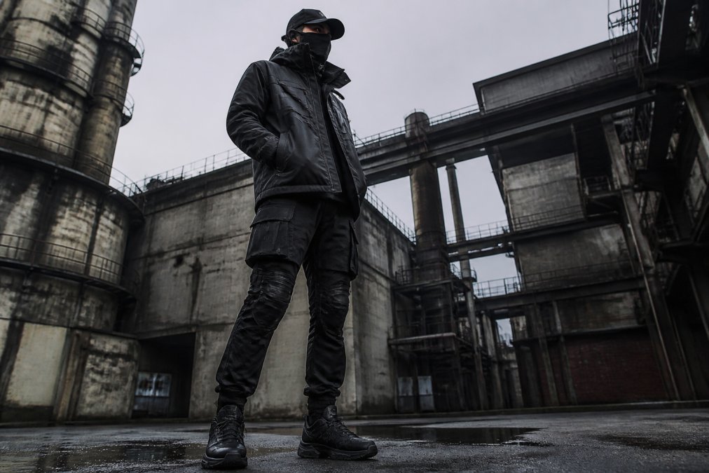Person wearing techwear outfit with fitted silhouette, articulated knee panels, and modular cargo pockets standing in urban environment with industrial architecture in background